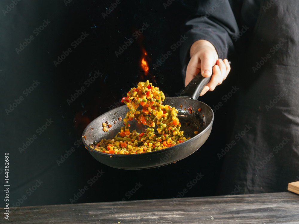 Cooking of vegetable mix stew with tossing on a hot frying pan in the tongues of flame. Colorful grains of corn and peas, asparagus and sweet peppers. Cooking of vegetable mix stew with tossing on a hot frying pan in the tongues of flame. Colorful grains of corn and peas, asparagus and sweet peppers.