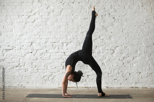 Young yogi attractive woman practicing yoga concept, standing in One legged Wheel exercise, Bridge pose, working out, wearing sportswear, black tank top and pants, full length, white loft background