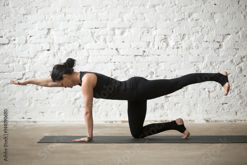 Young yogi attractive woman practicing yoga concept, standing in Donkey, Kick exercise, Bird dog pose, working out, wearing sportswear, black tank top and pants, full length, white loft background