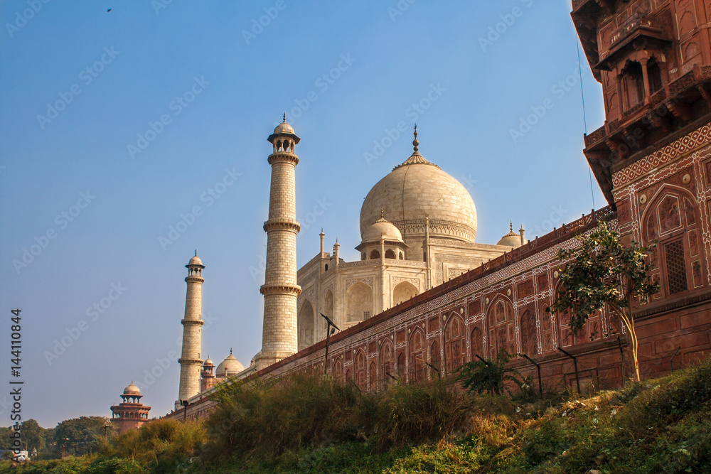 Taj Mahal in sun light with a little indian temple in the front. Early in the morning, back view behind the fence, from outside, river side. Image with a blue sky. One of the most famous building in
