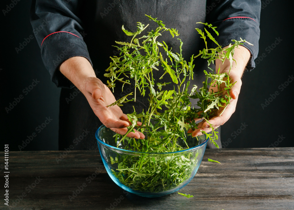 Mixing of a salad from fresh arugula in a glass bowl, standing on a black table top made of a textured wood board.