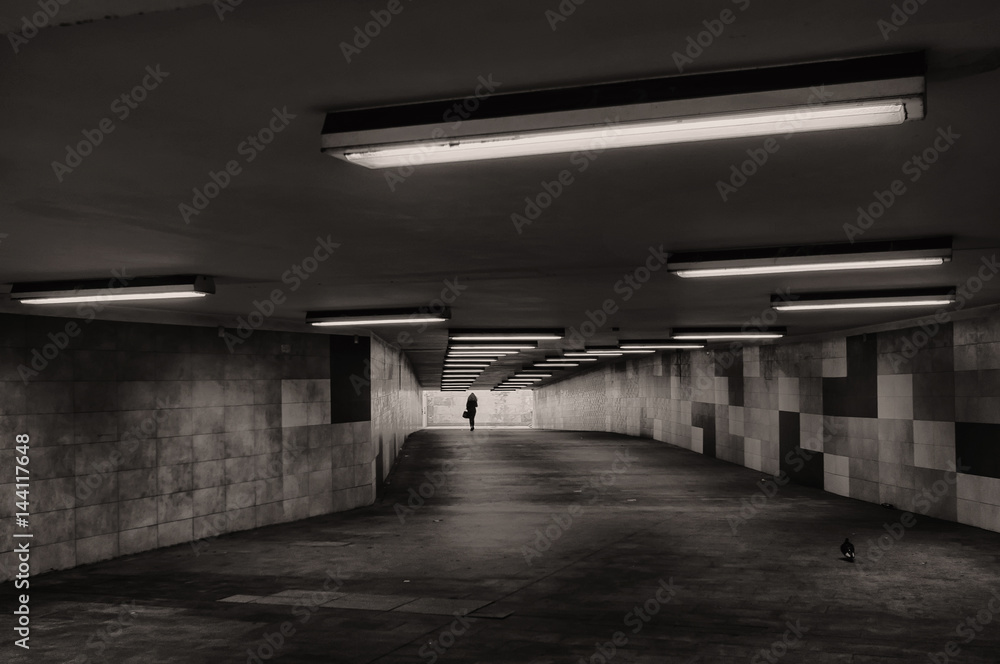 Black and white image of an underground passage. In the background, the silhouette of a woman is visible.