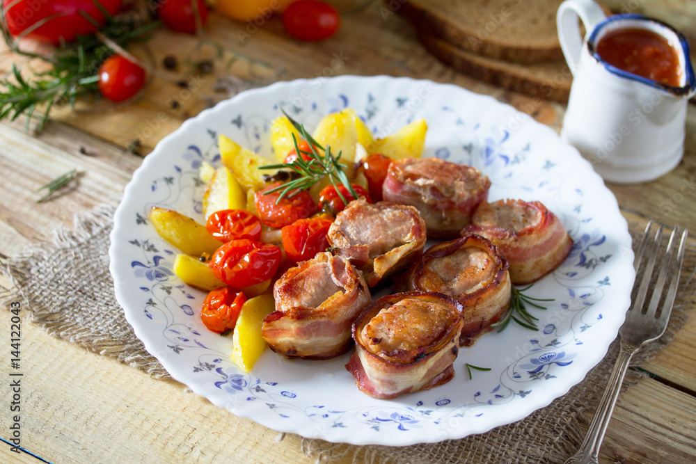 Pork fillet baked in bacon and baked potatoes, fresh vegetables and herbs on a wooden table in a rustic style.