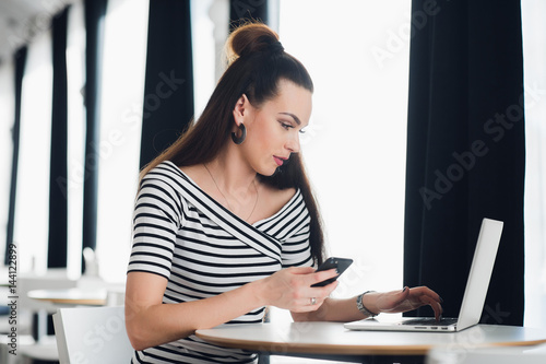 Portrait of beautiful businesswoman holding a phone while enjoying hot coffee in the cafe. Adult attractive female sitting at the table with a laptop and discussing work issues.