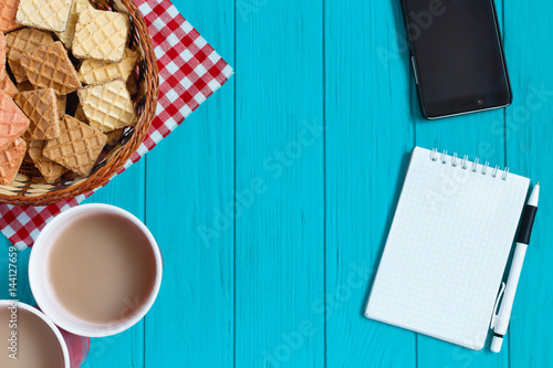 Bright turquoise table with tablet PC with blank screen, blank notebook,coffee and wafers, served in basket