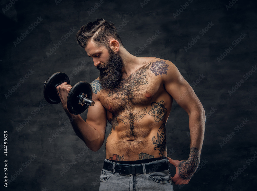 Shirtless bearded male with tattooed torso doing workout  with dumbbell.