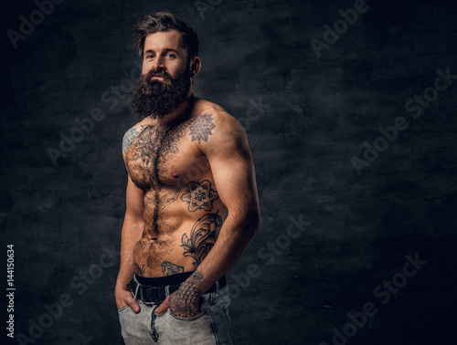 Bearded male with tattooed torso posing over dark grey background.