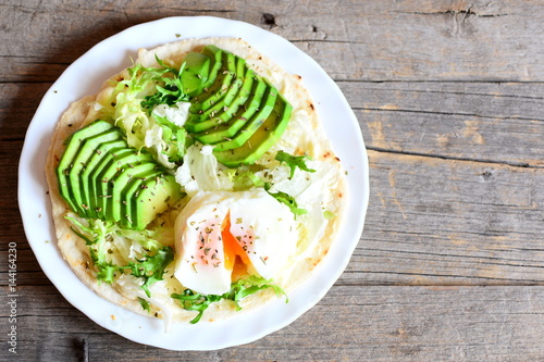 Poached egg, fresh avocado, salad, chinese cabbage, spices, sauce on a flour tortilla. Homemade tortilla with vegetarian filling on a plate isolated on old wooden background with copy space. Top view