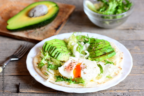 Healthy breakfast recipe. Homemade flour tortilla with a poached egg, avocado slices, napa cabbage, salad mix, sauce and spices on a plate. Avocado half, green salad in a glass bowl, fork on a table