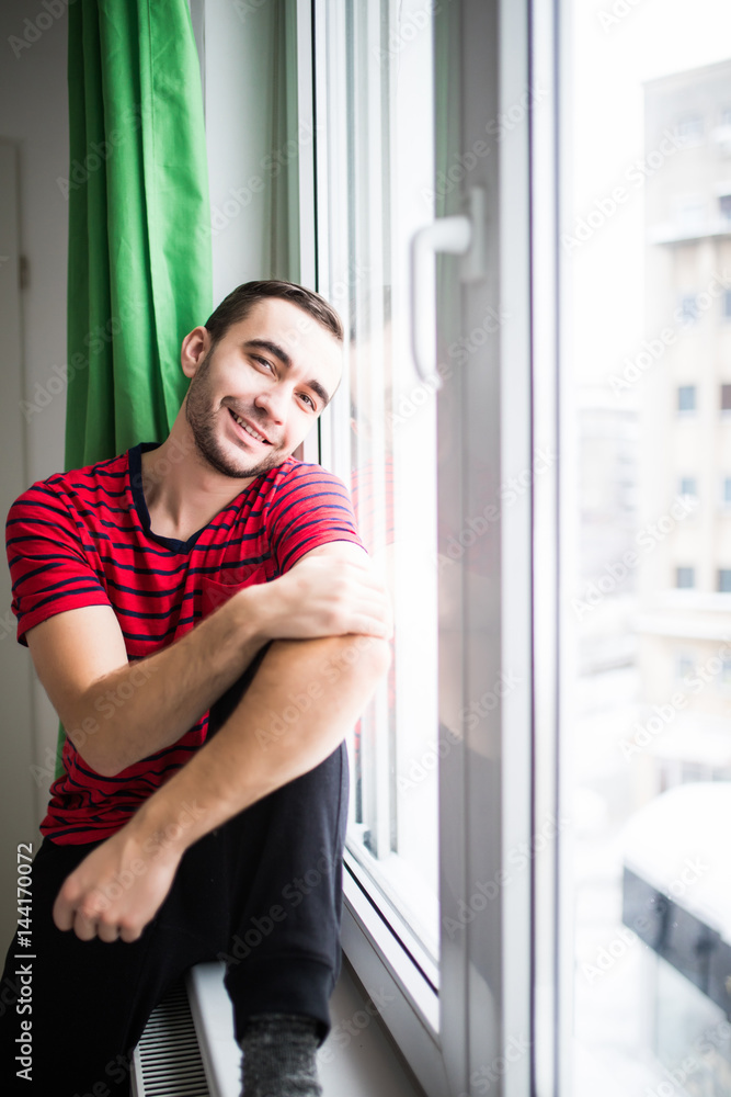 Young man smile to the camera in the morning sitting near window at his apparment Young man smile to the camera in the morning sitting near window at his apparment