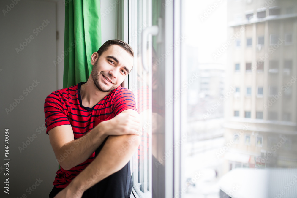 Young man smile to the camera in the morning sitting near window at his apparment Young man smile to the camera in the morning sitting near window at his apparment