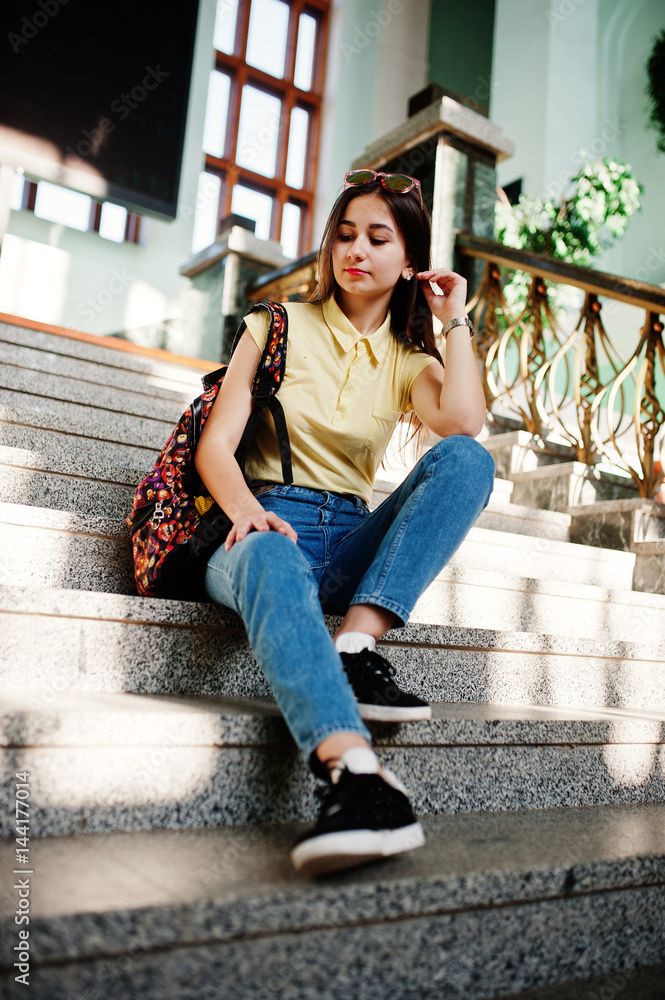 Young teenage girl sitting on stairs, wear on yellow t-shirt, jeans and sunglasses with backpack.