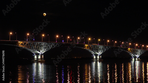 Full moon rises over the bridge. Road bridge between the cities of Saratov and Engels, Russia. The Volga River. The evening lights of cars and street lights