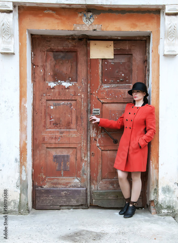 Young beautiful girl dressed in red coat posing near an old door from the entrance of the house. Female face in shadow. © Dmitriy