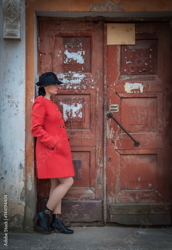 Young beautiful girl dressed in red coat posing near an old door from the entrance of the house. Female face in shadow. © Dmitriy