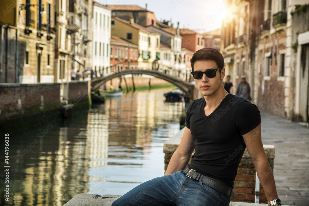 Portrait of Attractive Dark Haired Young Man Leaning Against Railing on Foot Bridge Over Narrow Canal in Venice, Italy