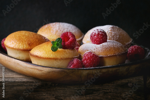 Homemade lemon muffin cupcakes with fresh raspberries, sugar powder, mint, served in square wood plate on wooden table over dark background. Dark rustic style. Close up