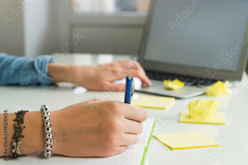 Female hands writing ideas on paper and working with laptop computer. Woman freelancer working at home office desk with notepad and yellow sticky notes