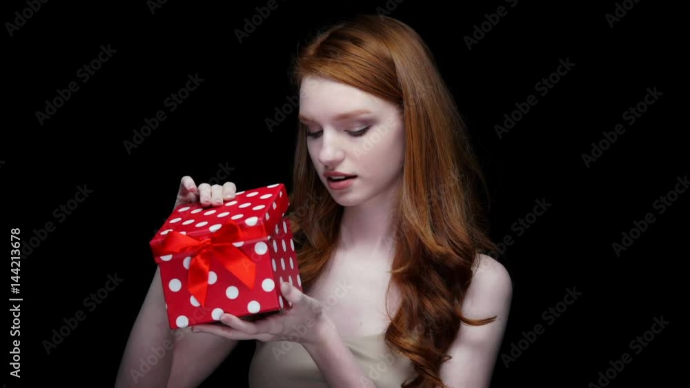 Disappointed upset redhead young woman opening gift box and looking at camera isolated on a black background Disappointed upset redhead young woman opening gift box and looking at camera isolated on a black background