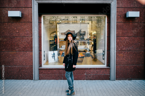 Portrait of beautiful and lovely model with long hair in fashion clothes, black sunglasses and elegant hat standing in front of boutique showcase in spring  and posing for camera. Celebrity style. © benevolente