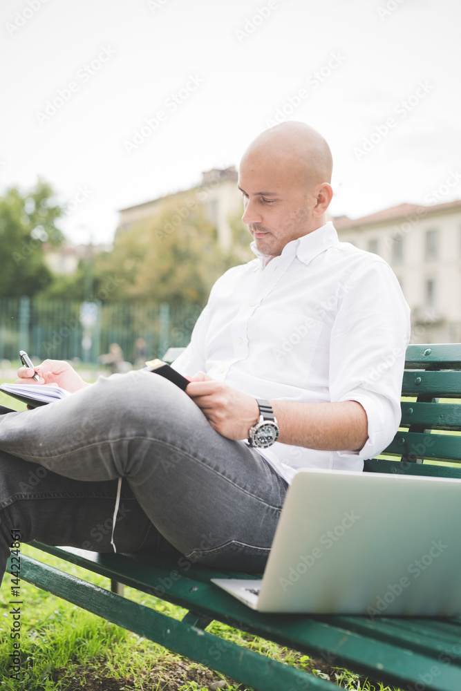 Fototapeta premium Young contemporary businessman remote working sitting on a bench outdoor in a city wiritng on her agenda - organizing, small business, remote working