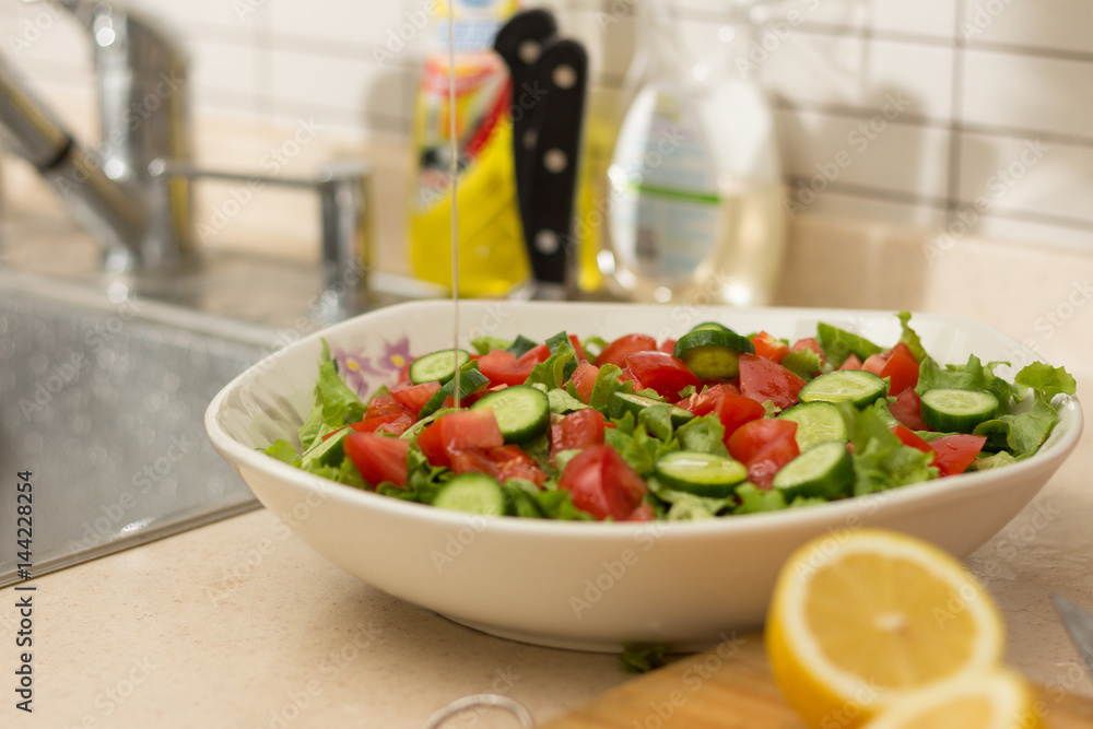Fresh salad with lettuce, cucumber and tomatoes in white big plate pouring olive oil and also lemon slices in fore ground in kitchen Fresh salad with lettuce, cucumber and tomatoes in white big plate pouring olive oil and also lemon slices in fore ground in kitchen