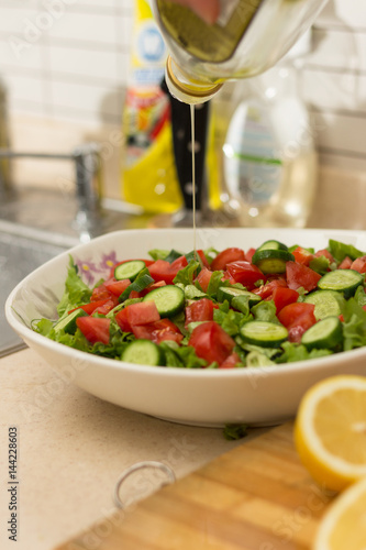 Fresh salad with lettuce, cucumber and tomatoes in white big plate and lemon slicesand knife on wooden chopping board on foreground in kitchen