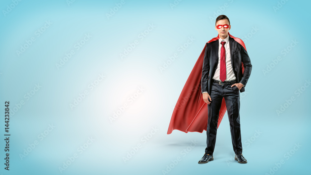 A self-assured businessman in a red cape and a mask standing with a hand in his pocket on blue background.