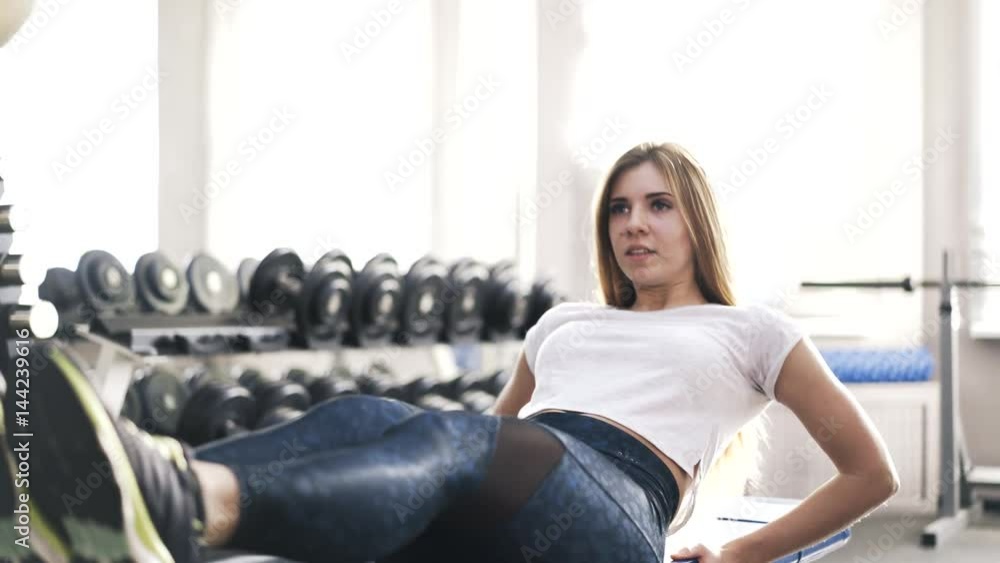 Young blond woman wearing a white T-shirt is doing abs exercise on a gym bench. Handheld real time close up shot Young blond woman wearing a white T-shirt is doing abs exercise on a gym bench. Handheld real time close up shot