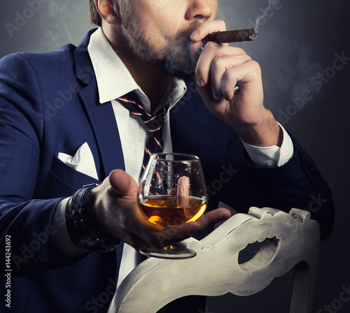 Cognac glass and cigar in a hand of a man dressed in suit and white shirt. Toned photo