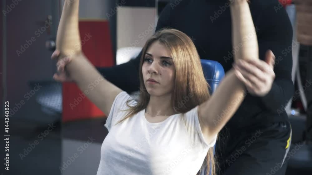 Close up of a young beautiful woman wearing a white T-shirt doing dumbbell exercise with a coach behind her. Tilt down real time close up shot Close up of a young beautiful woman wearing a white T-shirt doing dumbbell exercise with a coach behind her. Tilt down real time close up shot