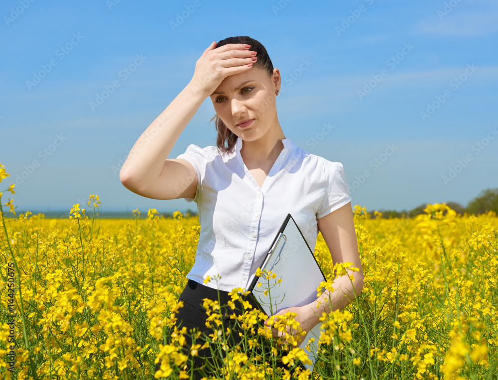 Sad business woman in flower field outdoor with clipboard touch head. Young girl in yellow rapeseed field. Beautiful spring landscape, bright sunny day Sad business woman in flower field outdoor with clipboard touch head. Young girl in yellow rapeseed field. Beautiful spring landscape, bright sunny day