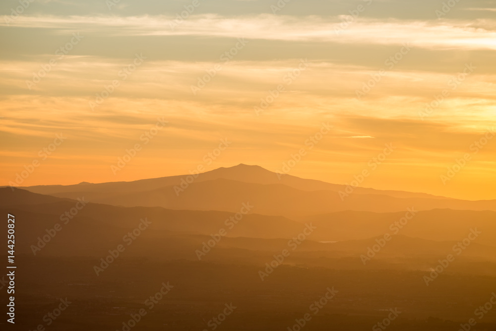 A mountain and some hills in the middle of a golden, orange light during sunset, with clouds also reflecting light