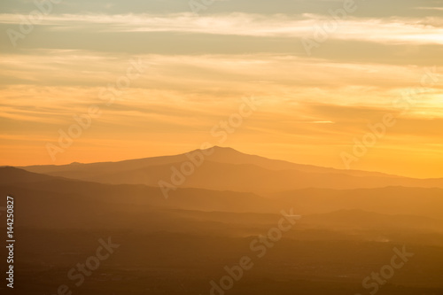 A mountain and some hills in the middle of a golden  orange light during sunset  with clouds also reflecting light