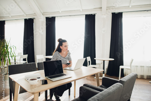 Adult woman holding mobile phone and typing messages while sitting at wooden table with open laptop computer.