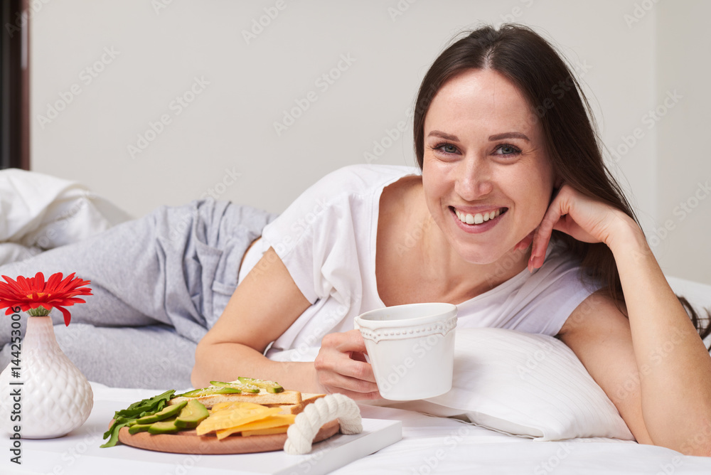 Beautiful brunette with long hair holding cup of tea while lying in bed