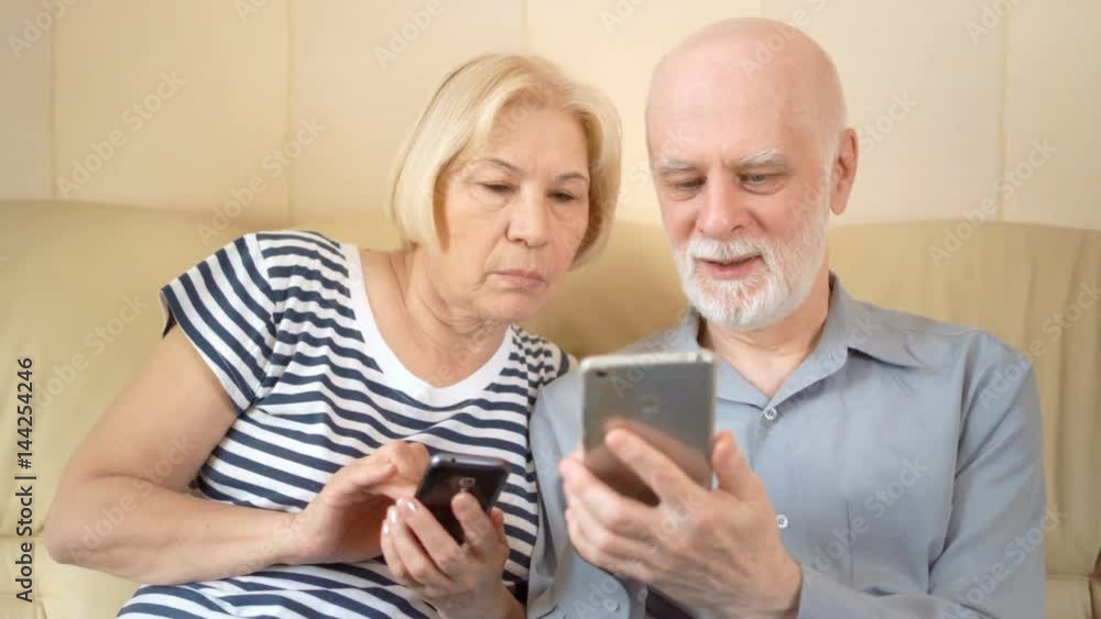 Cheerful good-looking senior couple sitting on sofa at home. Using smartphone, browsing, reading Cheerful good-looking senior couple sitting on sofa at home. Using smartphone, browsing, reading