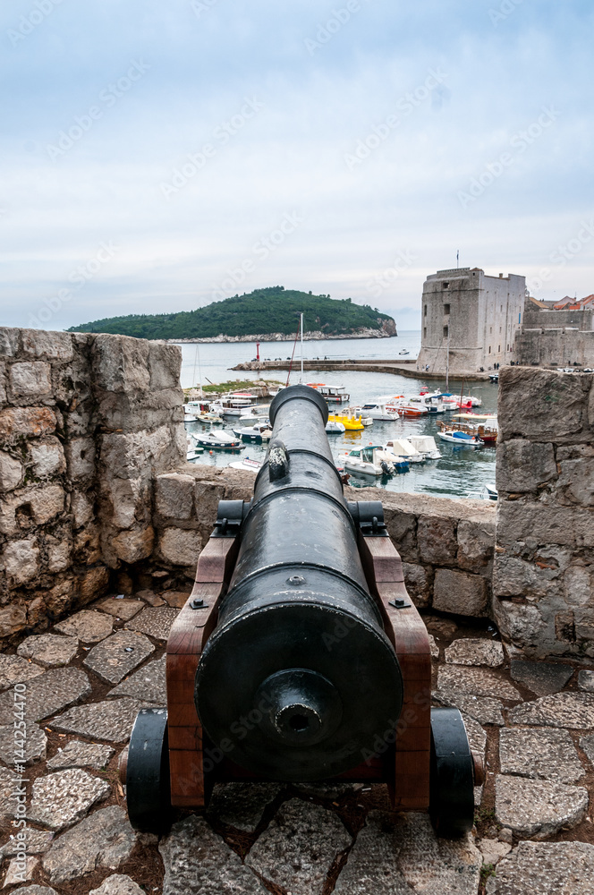 Large cannon jutting out from a fortified castellation, pointing over a coastal town.