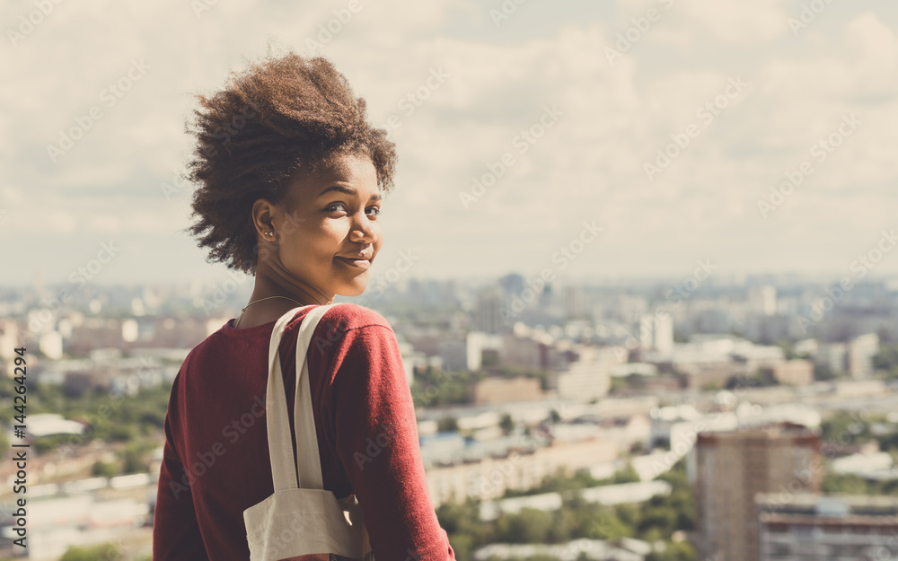 Portrait of young beautiful cute mixed Brazilian teen girl with curly afro hair standing on observation point very high and half-turned to camera, sunny summer cityscape below in blurred background