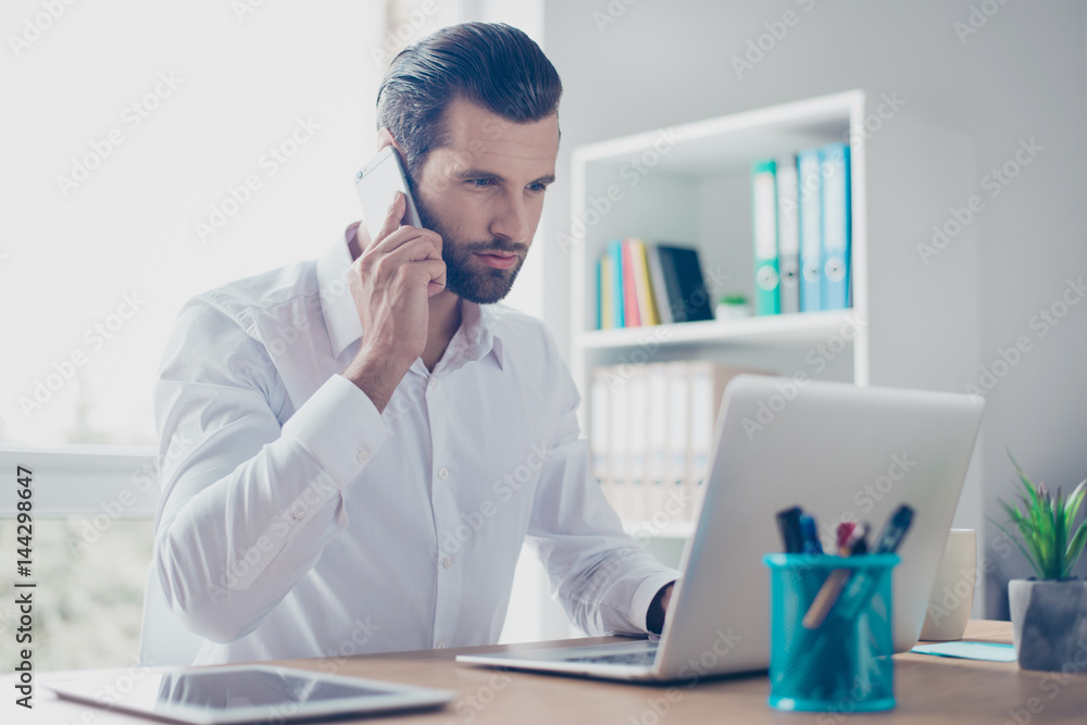 Concentrated and confident bearded businessman in white shirt talk on the phone while works on laptop Concentrated and confident bearded businessman in white shirt talk on the phone while works on laptop