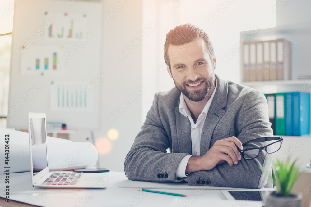 Confident smiling architect in formal wear holds glasses and sitting at the table in office Confident smiling architect in formal wear holds glasses and sitting at the table in office