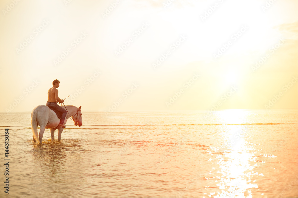 Fototapeta premium Macho man and horse on the background of sky and water. Boy model, cowboy on horseback on the beach by the sea at sunset. Men, backlit in sunshine. A positive summer time scene.