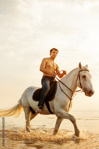 Fototapeta Naklejka Na Ścianę i Meble -  Macho man and horse on the background of sky and water. Boy model, cowboy on horseback on the beach by the sea at sunset. Men, backlit in sunshine. A positive summer time scene.