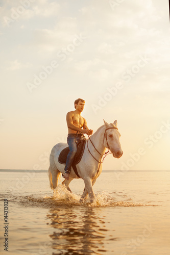 Macho man and horse on the background of sky and water. Boy model, cowboy on horseback on the beach by the sea at sunset. Men, backlit in sunshine. A positive summer time scene. © Miramiska