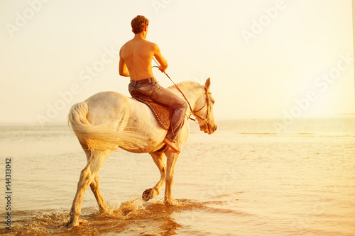 Macho man and horse on the background of sky and water. Boy model, cowboy on horseback on the beach by the sea at sunset. Men, backlit in sunshine. A positive summer time scene.