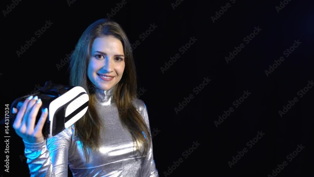 Cyber young woman in silver clothing showing virtual reality googles and gesturing thumb up, over dark background Cyber young woman in silver clothing showing virtual reality googles and gesturing thumb up, over dark background
