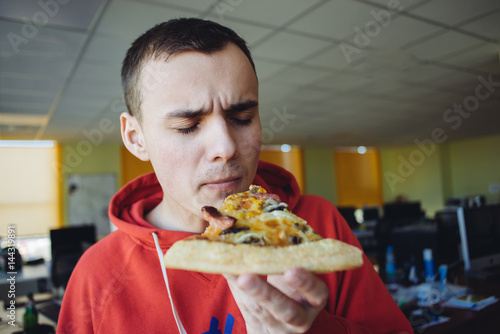 Portrait of a young man holding a large slice of pizza on a background of office.