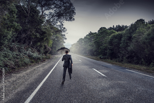 Back view of businessman with umbrella and suitcase walking on road . Mixed media