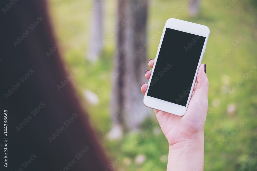 Mockup image of a woman using smart phone with blank black screen at outdoor and green nature background