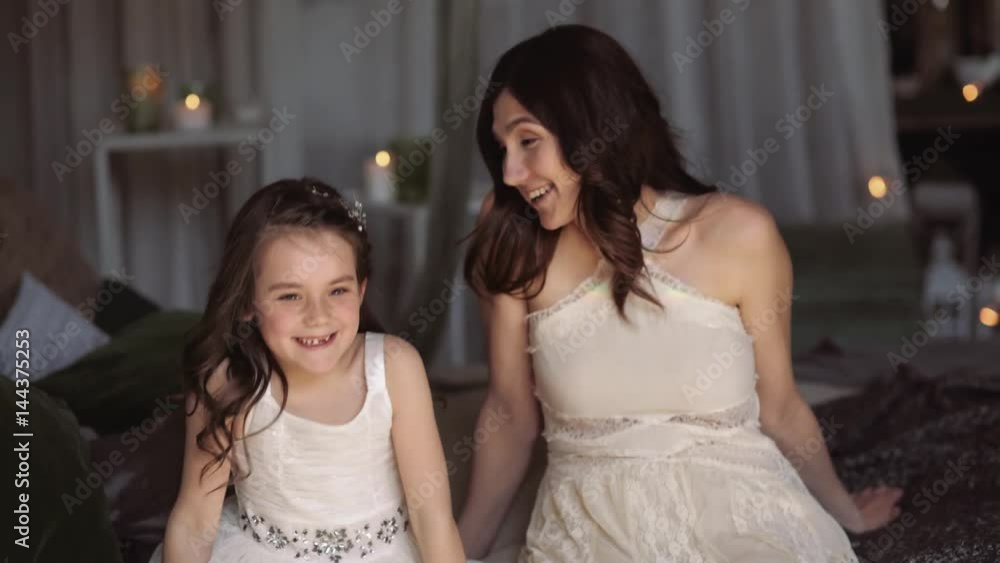 Mother and daughter hugging and cuddling in bed. Pretty little girl and beautiful woman lie together. Girls in lace dresses playing in decorated room. Family weekend, beauty day, having fun, love. Mother and daughter hugging and cuddling in bed. Pretty little girl and beautiful woman lie together. Girls in lace dresses playing in decorated room. Family weekend, beauty day, having fun, love.